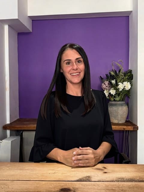 Woman with long dark hair in a black blouse sits at a wooden table, smiling, with a purple wall and potted flowers behind her.