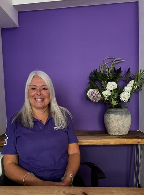 Smiling woman with long gray hair wearing a purple polo shirt sits beside a wooden shelf with a stone vase of white flowers against a purple wall.